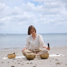Woman playing singing bowls on a beach with ocean and sky in the background